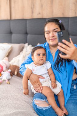 Rahatlamış bir anne bebeğini kucağında tutuyor. Akıllı telefonuyla selfie çekiyor. Günlük aile anlarını yakalıyor..