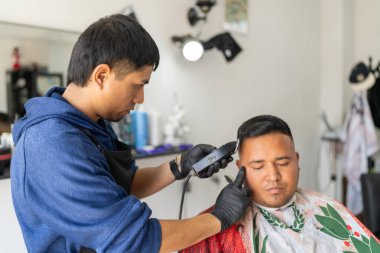 Genç Latin berber bir müşterinin saçını kesmek için elektrikli jilet kullanıyor.