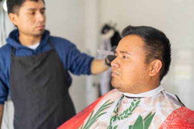 Üniformalı Latin genç berber bir müşterinin saçını tarıyor.