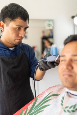 Bir müşterinin saçını kesmek için elektrikli tıraş makinesi kullanan Latin bir kuaförün dikey fotoğrafı.