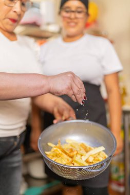 Bir aşçının ellerine odaklanmış dikey fotoğraf bir restoranın mutfağında kızarmış patatese tuz ekliyor.