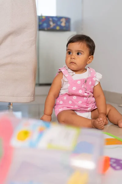 An adorable baby girl sits on the floor, her curious eyes gazing at something off-camera, with colorful toys nearby.