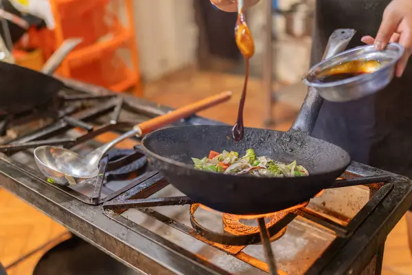 Kızartma tavasında karışık sebzelere sos ekleyen bir şefin yakın plan fotoğrafı.