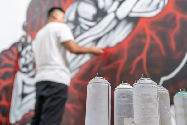 Low angle view photo with focus on several sprays on the floor while a muralist painting a wall