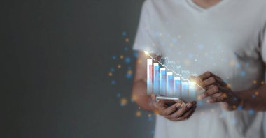 Genç adam para büyümesini görselleştirmek için akıllı telefon kullanıyor. Telefon ekranında artan yatırım ve tasarruf eğilimlerini gösteren grafikler ve çizelgeler ve artan bir hesap bakiyesi görülüyor.