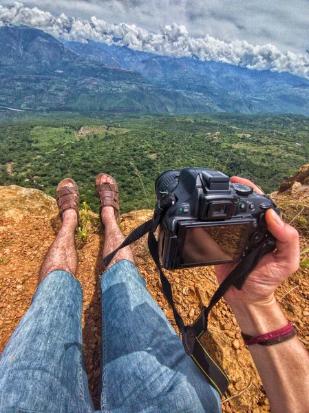 Maceraperest ve kaşif, kamerayla fotoğraf çekiyor. Bacakları ve ayakları kahverengi sandaletlerle ve mavi kot şortla. Yeşil orman ve turuncu kayalarla bir dağın zirvesinden manzara.