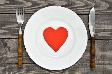 Sevgililer Günü sofrası. Beyaz tabakta kırmızı kalp sembolü ve ahşap masada çatal bıçak. 