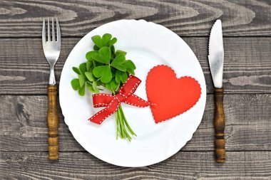 Valentins Günü masa takımı. Yeşil yonca yaprakları, beyaz tabakta kırmızı kalp sembolü ve ahşap masada kap kacak. 