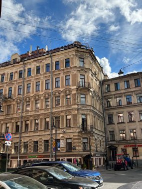 Parlak mavi gökyüzünün altında dağınık bulutlarla karışık mimari detayları olan tarihi bir Avrupa binası. Aşağıdaki cadde park halindeki araba ve yayalarla dolu. Eski bir şehir ortamının cazibesini yakalıyorlar..