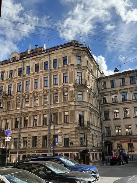 Parlak mavi gökyüzünün altında dağınık bulutlarla karışık mimari detayları olan tarihi bir Avrupa binası. Aşağıdaki cadde park halindeki araba ve yayalarla dolu. Eski bir şehir ortamının cazibesini yakalıyorlar..