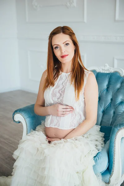 red-haired pregnant young girl in a blue armchair of a bright room