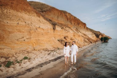 Deniz kıyısında, kil kayalıkların yanında beyaz elbiseli bir kızla bir adam.