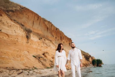 Deniz kıyısında, kil kayalıkların yanında beyaz elbiseli bir kızla bir adam.