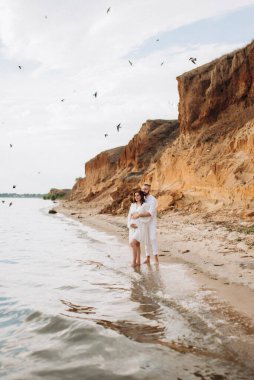 Deniz kıyısında, kil kayalıkların yanında beyaz elbiseli bir kızla bir adam.