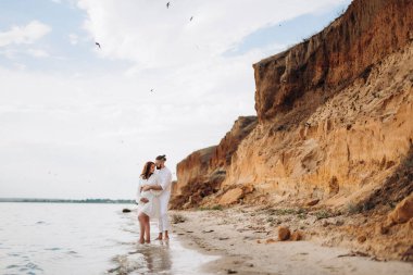 Deniz kıyısında, kil kayalıkların yanında beyaz elbiseli bir kızla bir adam.