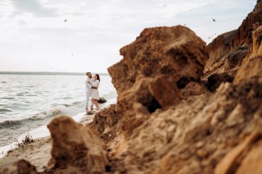 Deniz kıyısında, kil kayalıkların yanında beyaz elbiseli bir kızla bir adam.