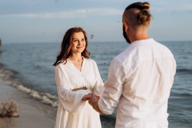 Deniz kıyısında, kil kayalıkların yanında beyaz elbiseli bir kızla bir adam.