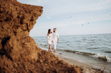 Deniz kıyısında, kil kayalıkların yanında beyaz elbiseli bir kızla bir adam.