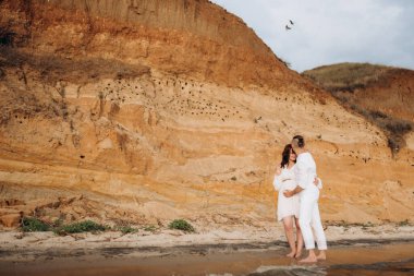 Deniz kıyısında, kil kayalıkların yanında beyaz elbiseli bir kızla bir adam.