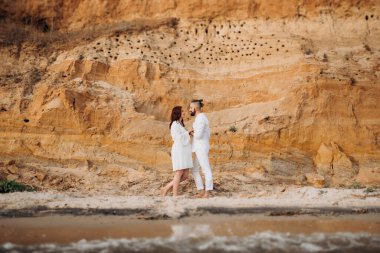 Deniz kıyısında, kil kayalıkların yanında beyaz elbiseli bir kızla bir adam.