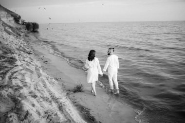 Deniz kıyısında, kil kayalıkların yanında beyaz elbiseli bir kızla bir adam.