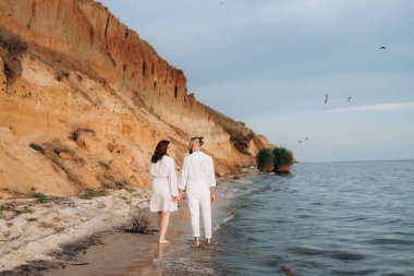 Deniz kıyısında, kil kayalıkların yanında beyaz elbiseli bir kızla bir adam.