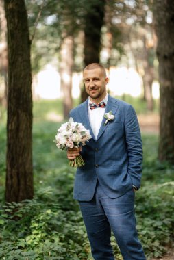 portrait of an elegant groom in a blue suit in a deciduous forest with a bouquet