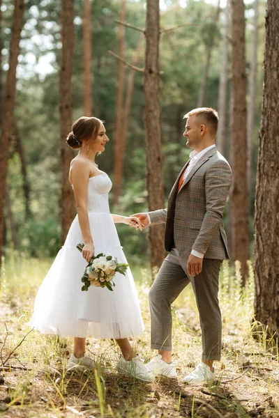 白いショートドレスを着た若いカップルの花嫁と松の森の中の灰色のスーツを着た新郎 — ストック写真