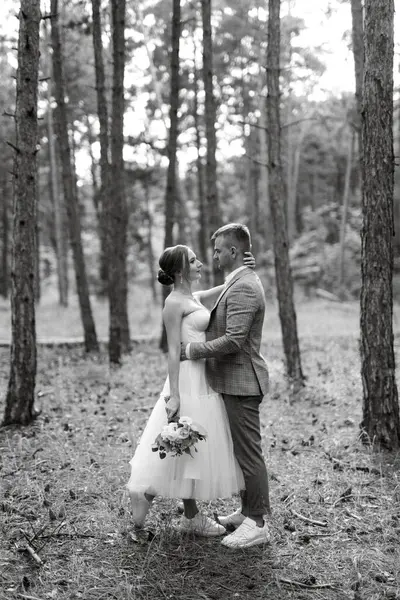 白いショートドレスを着た若いカップルの花嫁と松の森の中の灰色のスーツを着た新郎 — ストック写真