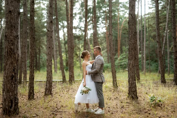 穿着白色短裙的年轻夫妇新娘 穿着灰色西装的新郎 在树林中的松树丛中 — 图库照片