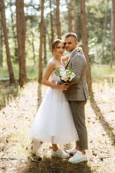 穿着白色短裙的年轻夫妇新娘 穿着灰色西装的新郎 在树林中的松树丛中 — 图库照片