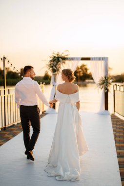 Gelin ve damat, nehrin kıyısındaki bir iskelede sarı bir günbatımının arkaplanına karşı.