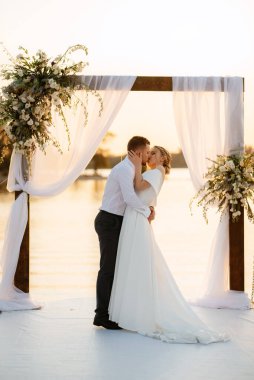 Gelin ve damat, nehrin kıyısındaki bir iskelede sarı bir günbatımının arkaplanına karşı.