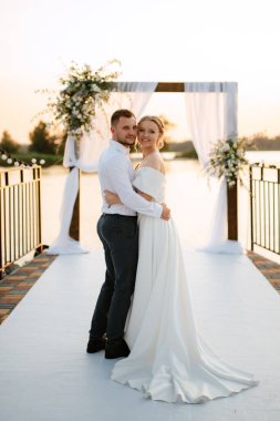 bride and groom against the backdrop of a yellow sunset on a pier near the river