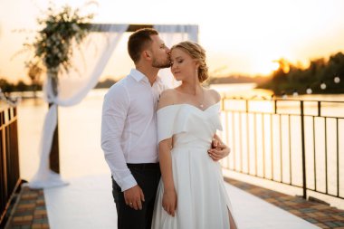 Gelin ve damat, nehrin kıyısındaki bir iskelede sarı bir günbatımının arkaplanına karşı.