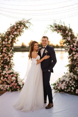 Gelin ve damat, nehrin kıyısındaki bir iskelede sarı bir günbatımının arkaplanına karşı.