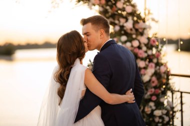 Gelin ve damat, nehrin kıyısındaki bir iskelede sarı bir günbatımının arkaplanına karşı.