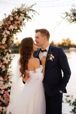 Gelin ve damat, nehrin kıyısındaki bir iskelede sarı bir günbatımının arkaplanına karşı.