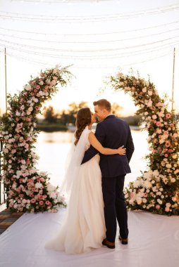 Gelin ve damat, nehrin kıyısındaki bir iskelede sarı bir günbatımının arkaplanına karşı.