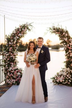 Gelin ve damat, nehrin kıyısındaki bir iskelede sarı bir günbatımının arkaplanına karşı.