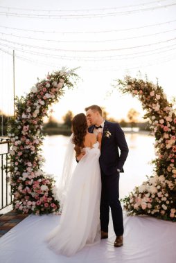 Gelin ve damat, nehrin kıyısındaki bir iskelede sarı bir günbatımının arkaplanına karşı.