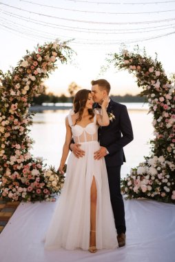 Gelin ve damat, nehrin kıyısındaki bir iskelede sarı bir günbatımının arkaplanına karşı.