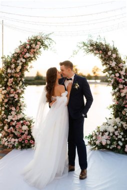 Gelin ve damat, nehrin kıyısındaki bir iskelede sarı bir günbatımının arkaplanına karşı.