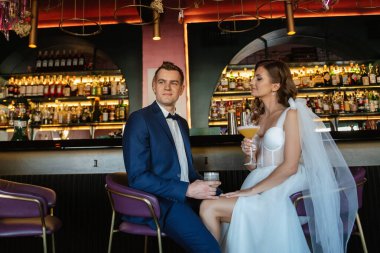 Gelin ve damat bir kokteyl barının içinde. Parlak bir atmosferde, bir bardak içkiyle.