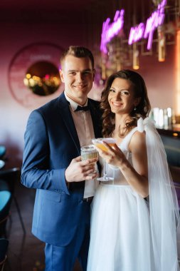 Gelin ve damat bir kokteyl barının içinde. Parlak bir atmosferde, bir bardak içkiyle.