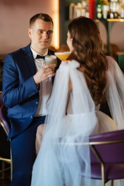 Gelin ve damat bir kokteyl barının içinde. Parlak bir atmosferde, bir bardak içkiyle.