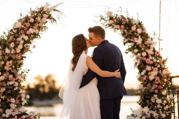 Gelin ve damat, nehrin kıyısındaki bir iskelede sarı bir günbatımının arkaplanına karşı.