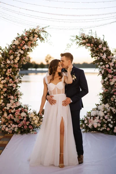 Gelin ve damat, nehrin kıyısındaki bir iskelede sarı bir günbatımının arkaplanına karşı.