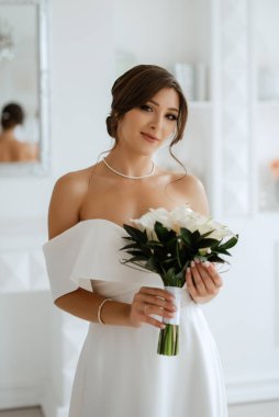 Parlak bir stüdyoda, dar bir gelinlik içinde, buketi olan esmer gelin.