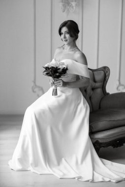 Dar bir gelinlik içinde, parlak bir stüdyoda, koltukta bir buketle esmer gelin.